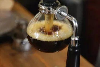 A steaming cup of hand-brewed coffee beside a hand brew pot, filter cup, and freshly ground coffee, showcasing the perfect home brewing setup.